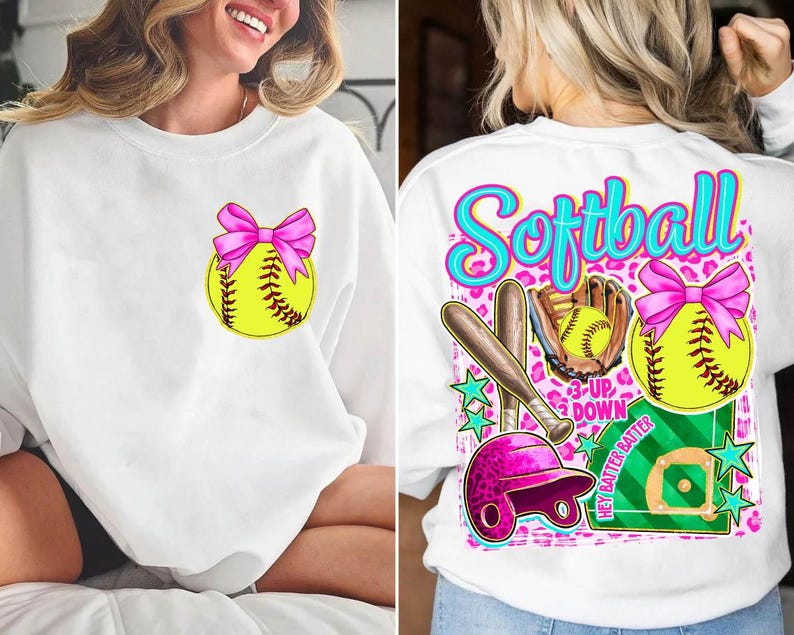 May include: White sweatshirt featuring a softball design. The front has a yellow softball with a pink bow. The back has a larger design with the word "Softball", a bat, glove, helmet, and a softball with a pink bow.