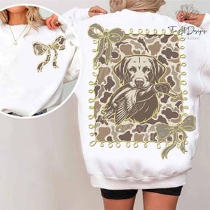 May include: White sweatshirt featuring a bow design on the front and back. The back design includes a dog holding a duck, set against a camouflage pattern, with a decorative border and bows. The design is in shades of brown and tan.