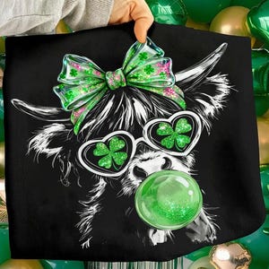 May include: Black graphic tee featuring a Highland cow with a green bow, heart-shaped sunglasses with shamrocks, and a green glitter ball. The cow's fur is white, and the sunglasses have a white frame. The bow has pink accents.