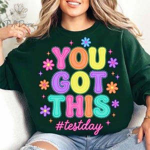 Pode incluir: Camisola verde escuro com a frase "YOU GOT THIS" em letras coloridas e arredondadas, decorada com flores e a hashtag "#testday". Uma pe&ccedil;a casual e confort&aacute;vel.