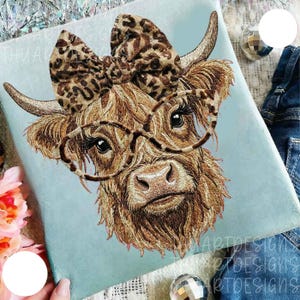Op de afbeelding: Lichtblauw T-shirt met een borduursel van een Schotse Hooglander koe met een bril en een luipaardprint strik. De koe heeft lang, ruig bruin haar en kijkt recht in de camera. De tekst "ARTDESIGNS" is zichtbaar.