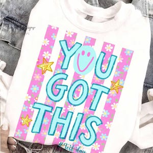 May include: White sweatshirt with the text "YOU GOT THIS" in turquoise, a smiley face, and gold star accents. The design includes pink vertical stripes with a floral pattern. The hashtag "#Test day" is at the bottom.