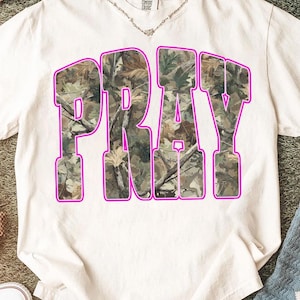 May include: White t-shirt with the word "PRAY" in large, block letters. The letters are filled with a camouflage pattern and outlined in bright pink. The shirt has a crew neck and short sleeves.