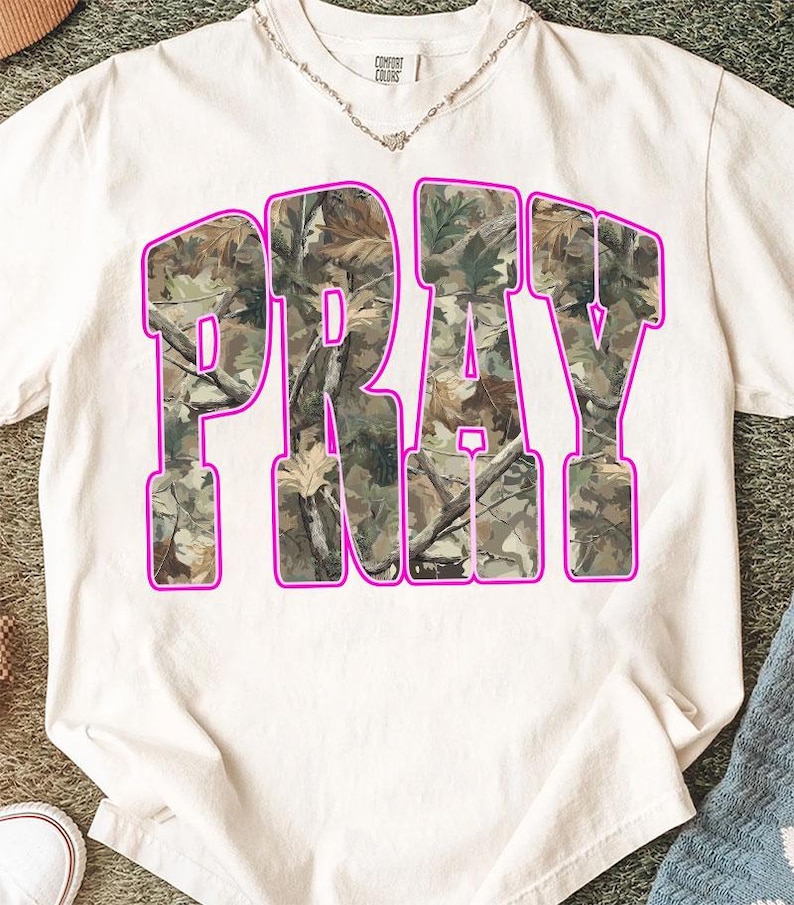 May include: White t-shirt with the word "PRAY" in large, block letters. The letters are filled with a camouflage pattern and outlined in bright pink. The shirt has a crew neck and short sleeves.