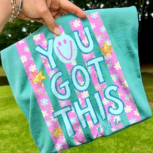 May include: Teal t-shirt with the phrase "YOU GOT THIS" in large, light blue letters. Pink stripes with floral patterns and gold stars accent the design. The hashtag "#Test day" is at the bottom.