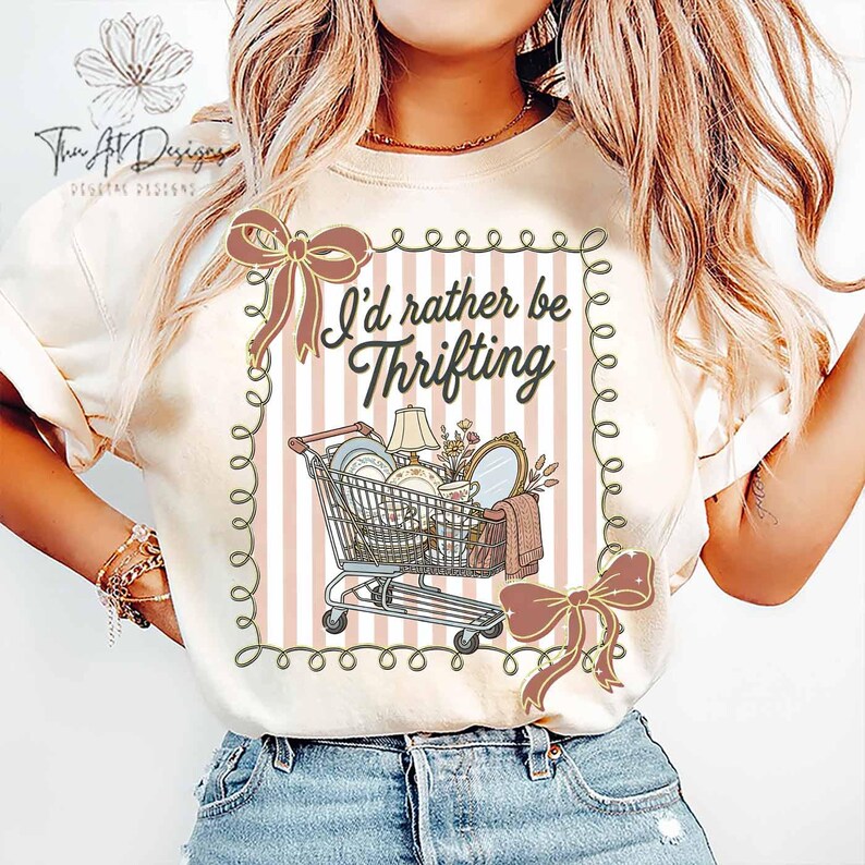 May include: Cream-colored t-shirt with the text "I'd rather be Thrifting" above a shopping cart graphic filled with vintage items. The design includes pink and white stripes, bows, and decorative swirls.