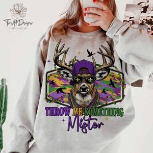 May include: A light gray sweatshirt featuring a deer wearing a purple cap, sunglasses, and Mardi Gras beads. The deer is set against a camouflage background with the text "THROW ME SOMETHING Mister" in purple and green.