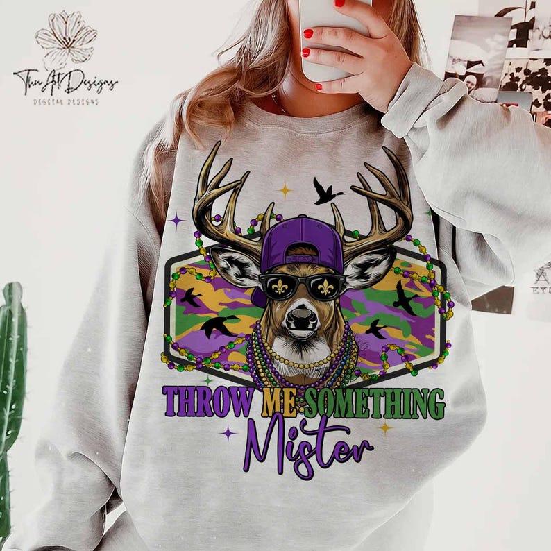 May include: A light gray sweatshirt featuring a deer wearing a purple cap, sunglasses, and Mardi Gras beads. The deer is set against a camouflage background with the text "THROW ME SOMETHING Mister" in purple and green.