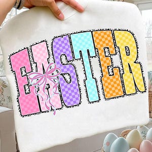 May include: A white sweatshirt featuring the word "EASTER" in colorful, checkered letters. The letters are outlined in black and accented with a pink bow. The sweatshirt is held up by a hand.