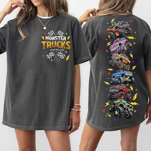Puede incluir: Camiseta gris oscuro con el texto "MONSTER TRUCKS ARE MY JAM" y banderas a cuadros. La parte trasera de la camiseta presenta una disposici&oacute;n vertical de ilustraciones de monster trucks coloridos con acentos de rayos y estrellas.