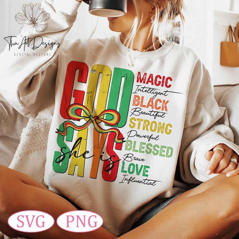 Op de afbeelding: Een wit sweatshirt met een kleurrijk grafisch ontwerp dat "God Says She Is" zegt met de woorden "Magic, Intelligent, Black, Beautiful, Strong, Powerful, Blessed, Brave, Love, Influential" verticaal aan de rechterkant van de grafiek vermeld.