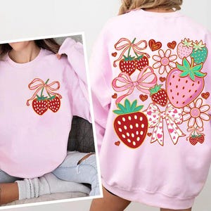 Könnte beinhalten: Rosafarbener Pullover mit einem Grafikdruck von Erdbeeren, Schleifen und Herzen. Die Erdbeeren sind rot mit weißen Kernen und grünen Blättern. Die Schleifen sind rosa mit weißen Punkten. Die Herzen sind rosa.