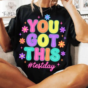 Pode incluir: T-shirt preto com a frase "YOU GOT THIS" em letras coloridas, decorado com flores e estrelas. A hashtag "#testday" est&aacute; escrita abaixo em rosa.