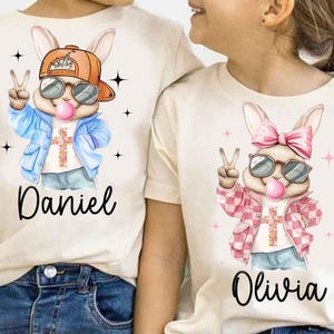 Può includere: Due magliette bianche con un coniglio dei cartoni animati che indossa occhiali da sole e un segno di pace. Il coniglio a sinistra indossa una giacca blu e ha il nome "Daniel" stampato sotto. Il coniglio a destra indossa una giacca a scacchi rosa e bianca e ha il nome "Olivia" stampato sotto.