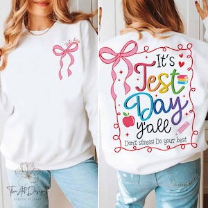 May include: White sweatshirt with a pink bow on the front and the text "It's Test Day Y'all Don't stress! Do your best" on the back. The text is in rainbow colors with a red apple, a pencil, and a stack of books.