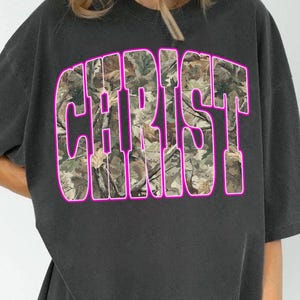 May include: Dark gray t-shirt featuring the word "CHRIST" in large, block letters. The letters are filled with a camouflage pattern and outlined in bright pink. The shirt has short sleeves and a crew neck.