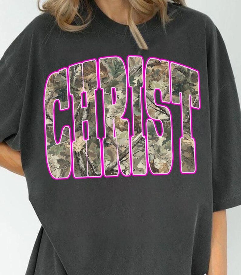 May include: Dark gray t-shirt featuring the word "CHRIST" in large, block letters. The letters are filled with a camouflage pattern and outlined in bright pink. The shirt has short sleeves and a crew neck.