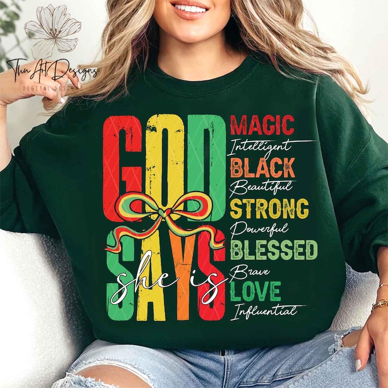Op de afbeelding: Groen sweatshirt met een kleurrijke afbeelding waarop staat "God Says she is" met de woorden "Magic, Intelligent, Black, Beautiful, Strong, Powerful, Blessed, Brave, Love, Influential" die verticaal aan de rechterkant van de afbeelding zijn gedrukt.
