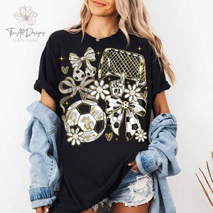 May include: Black t-shirt with a soccer-themed graphic. The design includes soccer balls, bows, flowers, a soccer goal, and the number one hand gesture. The text "Twin Art Designs" is visible in the upper left corner.