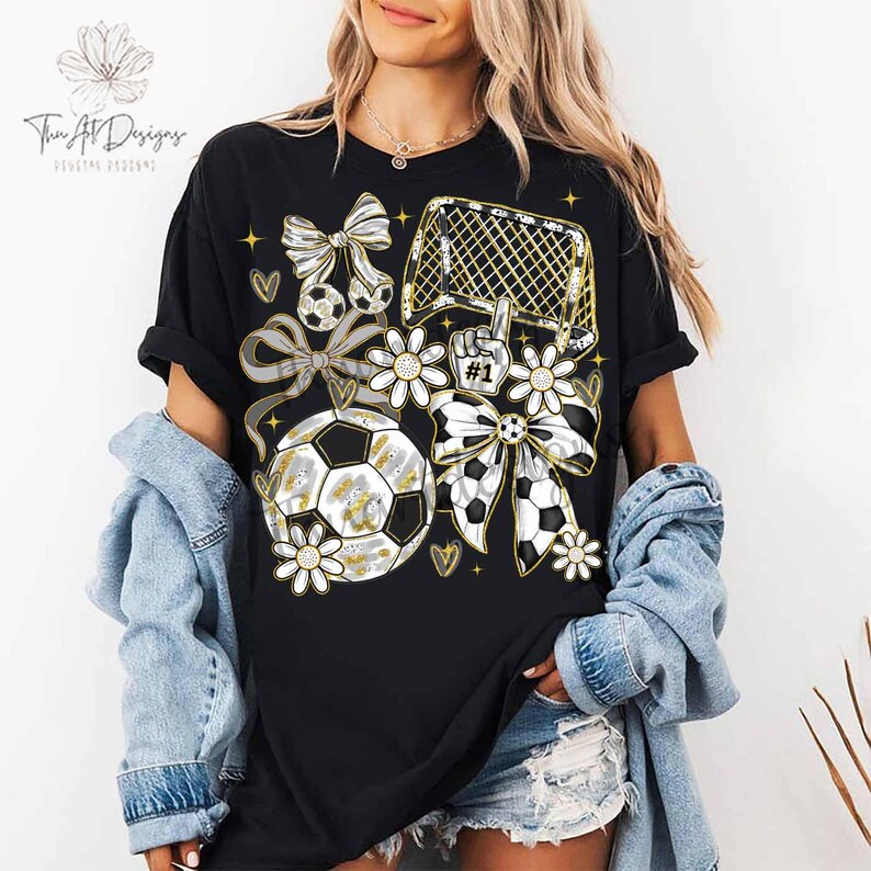 May include: Black t-shirt with a soccer-themed graphic. The design includes soccer balls, bows, flowers, a soccer goal, and the number one hand gesture. The text "Twin Art Designs" is visible in the upper left corner.