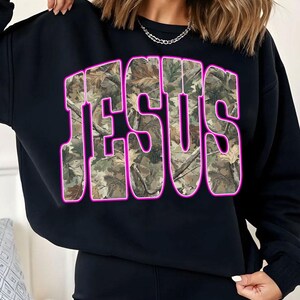 May include: Black sweatshirt with the word "JESUS" in large, bold letters across the front. The letters are filled with a camouflage pattern and outlined in bright pink.