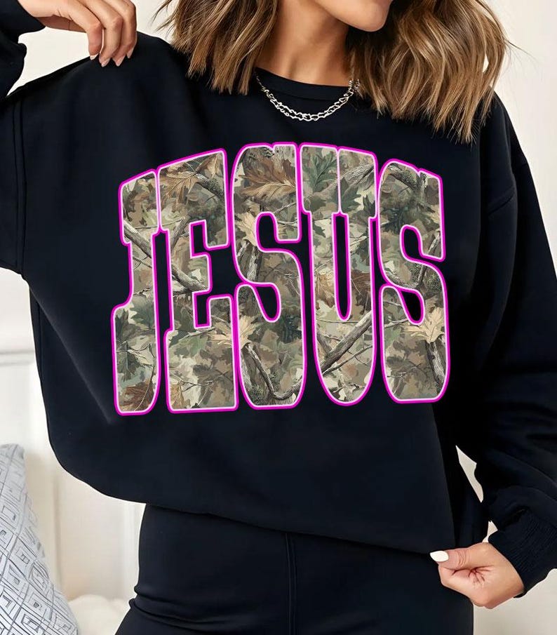 May include: Black sweatshirt with the word "JESUS" in large, bold letters across the front. The letters are filled with a camouflage pattern and outlined in bright pink.