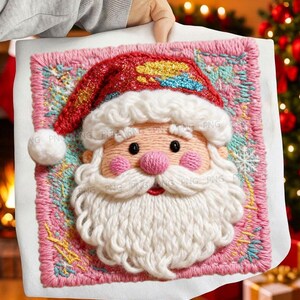 Könnte beinhalten: Besticktes Weihnachtskissen mit Weihnachtsmann. Das Gesicht des Weihnachtsmanns hat einen weißen Bart, rosa Wangen und einen roten Hut mit goldenen und blauen Akzenten. Das Kissen hat einen rosa Rand mit einem Schneeflockenmuster.