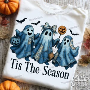 May include: White sweatshirt featuring three blue ghost characters with patchwork details, holding a jack-o'-lantern, a bow, and a pumpkin lollipop. The text "Tis The Season" is printed below the ghosts, with black bats in the background.