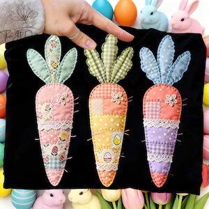 Op de afbeelding: Zwarte stof versierd met drie kleurrijke, patchwork wortel applicaties. Elke wortel heeft een ander patroon, waaronder bloemen, ruitjes en stippen, met kanten rand en bloemaccenten. Paas thema.