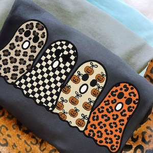 Peut inclure: Un sweat-shirt gris foncé avec quatre motifs de fantômes : imprimé léopard, damier noir et blanc, visages de citrouilles et imprimé léopard orange. Le sweat-shirt est plié avec d'autres tissus de couleur claire.