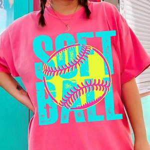 May include: A bright pink t-shirt featuring the word "SOFTBALL" in turquoise block letters. A yellow softball with pink stitching is in the centre. The design is bold and colourful.
