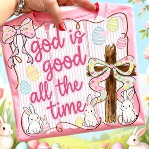Pode incluir: Camiseta rosa com a frase "god is good all the time" em rosa. O design inclui uma cruz de madeira, ovos de Páscoa, coelhos e laços. A camiseta é segurada por uma mão.