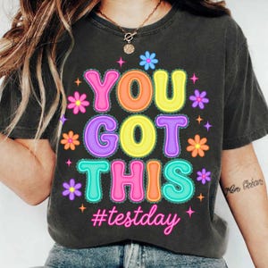 Pode incluir: T-shirt cinza escuro com a frase "YOU GOT THIS" em letras coloridas, rodeada por flores e estrelas. A hashtag "#testday" est&aacute; abaixo. A camisa &eacute; de estilo casual.