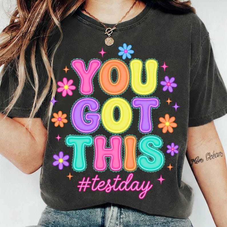 Pode incluir: T-shirt cinza escuro com a frase "YOU GOT THIS" em letras coloridas, rodeada por flores e estrelas. A hashtag "#testday" est&aacute; abaixo. A camisa &eacute; de estilo casual.