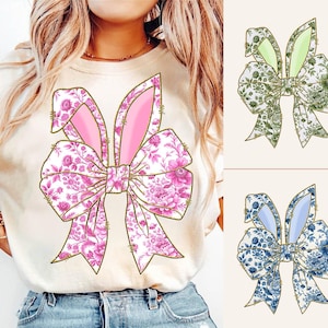 May include: A white t-shirt with a pink floral bunny bow design. The bow has pink bunny ears and is tied with a gold ribbon.