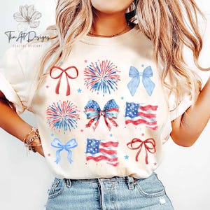 May include: A white t-shirt with a red, white, and blue design featuring fireworks, bows, and American flags. The design is a watercolor style.