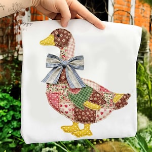 Può includere: Un articolo in tessuto bianco con un design a patchwork di anatra. L'anatra ha un becco e zampe gialle, una testa marrone e un corpo composto da vari tessuti fantasia in rosa, rosso, verde e marrone. Un fiocco blu e bianco adorna il collo.