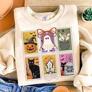 May include: Cream-colored sweatshirt featuring a Halloween-themed stamp design. The stamps depict a jack-o'-lantern, ghost, cauldron, black cat, haunted house, and a tombstone with "Trick or Treat" text. The design includes bats, a witch hat, and a bow.