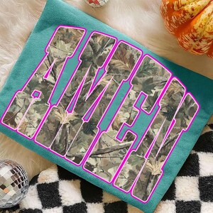 May include: Teal-colored t-shirt with the word "AMEN" in large, block letters. The letters are filled with a camouflage pattern and outlined in bright pink. The shirt is displayed on a fluffy white surface, with a disco ball and a pumpkin in the background.