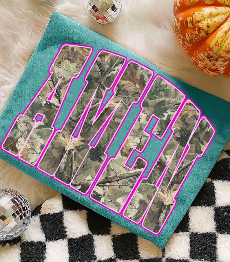 May include: Teal-colored t-shirt with the word "AMEN" in large, block letters. The letters are filled with a camouflage pattern and outlined in bright pink. The shirt is displayed on a fluffy white surface, with a disco ball and a pumpkin in the background.