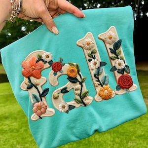 May include: A turquoise sweatshirt with the embroidered word "FALL" and the number "211". The letters and numbers are beige and decorated with colorful floral embroidery, including orange, red, and white flowers, and green leaves.