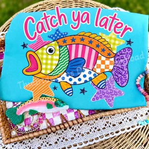 Puede incluir: Una camiseta de color turquesa brillante con un gráfico de pez colorido y estampado con el texto Catch ya later en rosa. El diseño del pez incluye secciones amarillas, verdes, naranjas y azules con varios patrones. Varias estrellas multicolores rodean al pez.