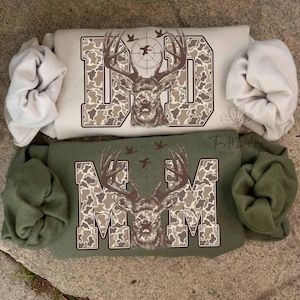 May include: Two sweatshirts, one tan and one green, each with a deer graphic. The word "DAD" is printed on the tan sweatshirt, and "MOM" is printed on the green sweatshirt. The deer graphic includes a target and flying birds.