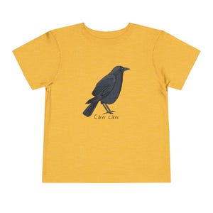 May include: A mustard yellow short-sleeve t-shirt featuring a graphic illustration of a black crow standing in profile. Below the bird, the text "Caw caw" is printed in a simple, dark brown font. The shirt has a crew neckline and a plain back.