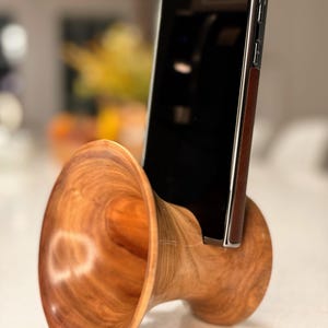 May include: A handcrafted wooden phone stand with a natural wood grain finish. The stand features a unique, horn-like design, holding a black smartphone with a brown leather case. The stand is designed to hold a phone in an upright position.