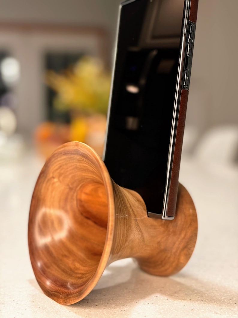 May include: A handcrafted wooden phone stand with a natural wood grain finish. The stand features a unique, horn-like design, holding a black smartphone with a brown leather case. The stand is designed to hold a phone in an upright position.