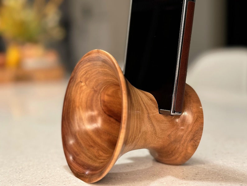 May include: A handcrafted wooden phone stand with a unique, horn-like design. The stand is made of polished, warm-toned wood, and holds a smartphone. The stand's design is both functional and decorative, adding a touch of natural elegance.