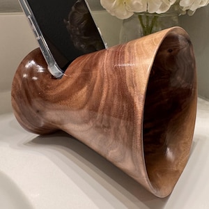 May include: A wooden phone stand with a curved design. The stand is made of dark brown wood with a natural grain pattern. A smartphone is placed in the stand.