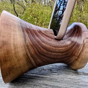 Op de afbeelding: Een houten telefoonstandaard gemaakt van walnoot. De standaard heeft een unieke vorm met een gebogen basis en een brede bovenkant die een telefoon rechtop houdt.