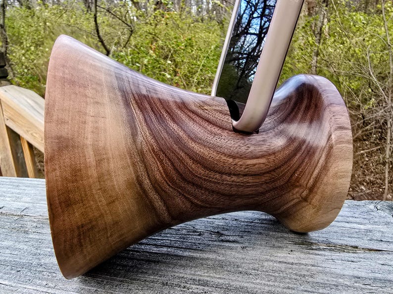 Op de afbeelding: Een houten telefoonstandaard gemaakt van walnoot. De standaard heeft een unieke vorm met een gebogen basis en een brede bovenkant die een telefoon rechtop houdt.
